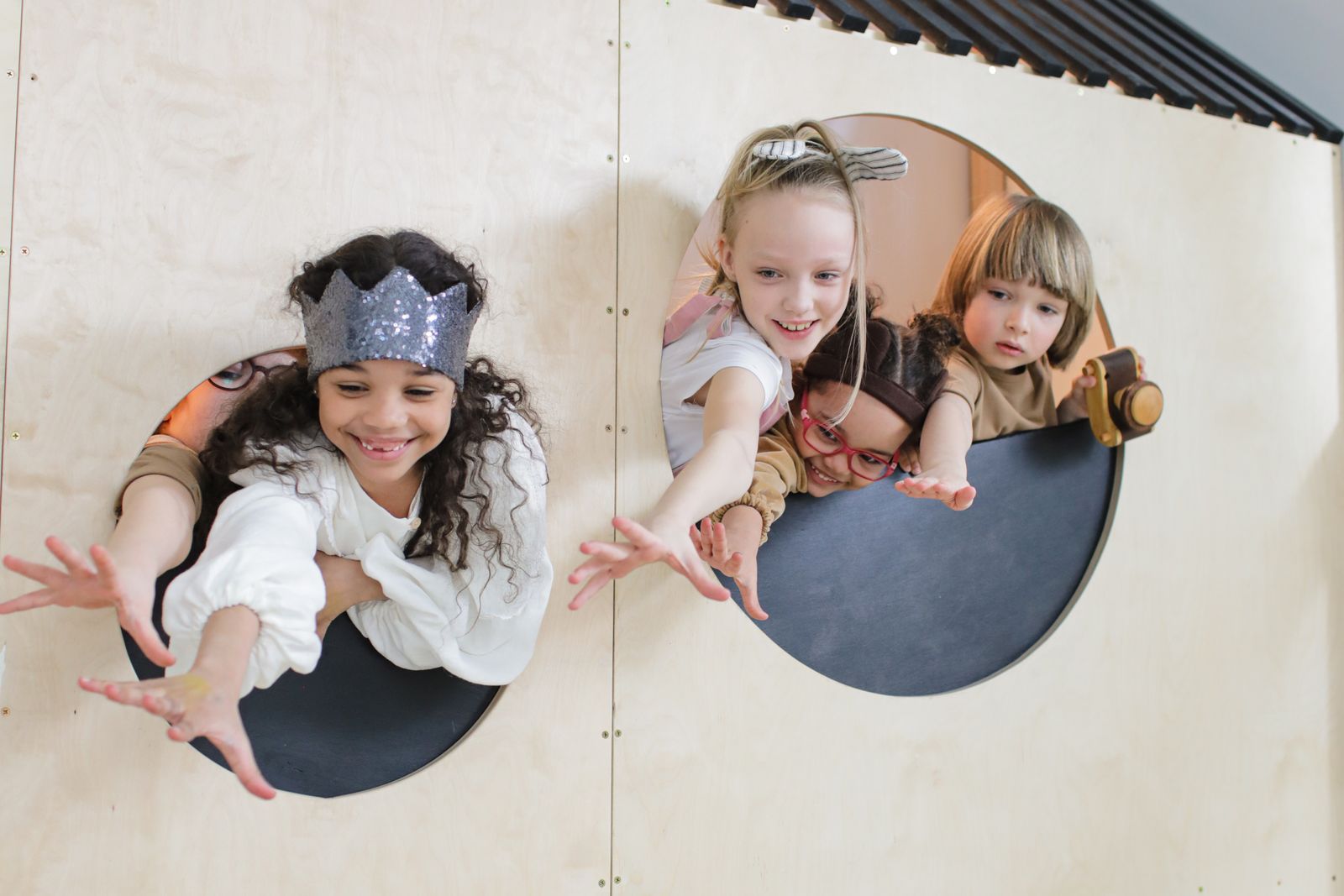 Four happy children reaching out and smiling through round porthole windows in a wooden play den
